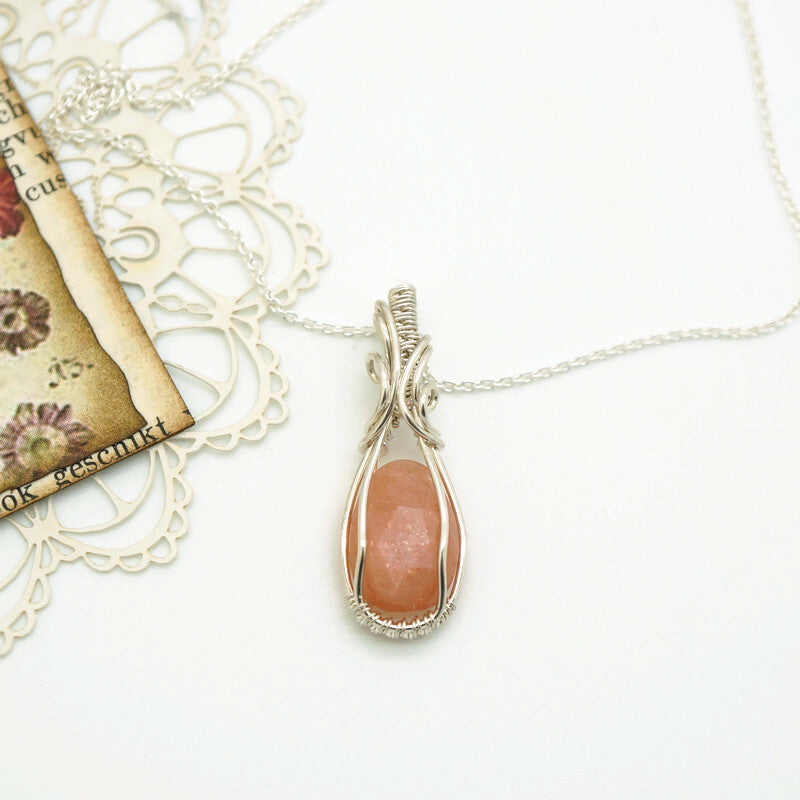 Unieke handgemaakte wire wrap hanger van zilver met perzikkleurige maansteen, liggend op witte ondergrond met vintage decoratie.