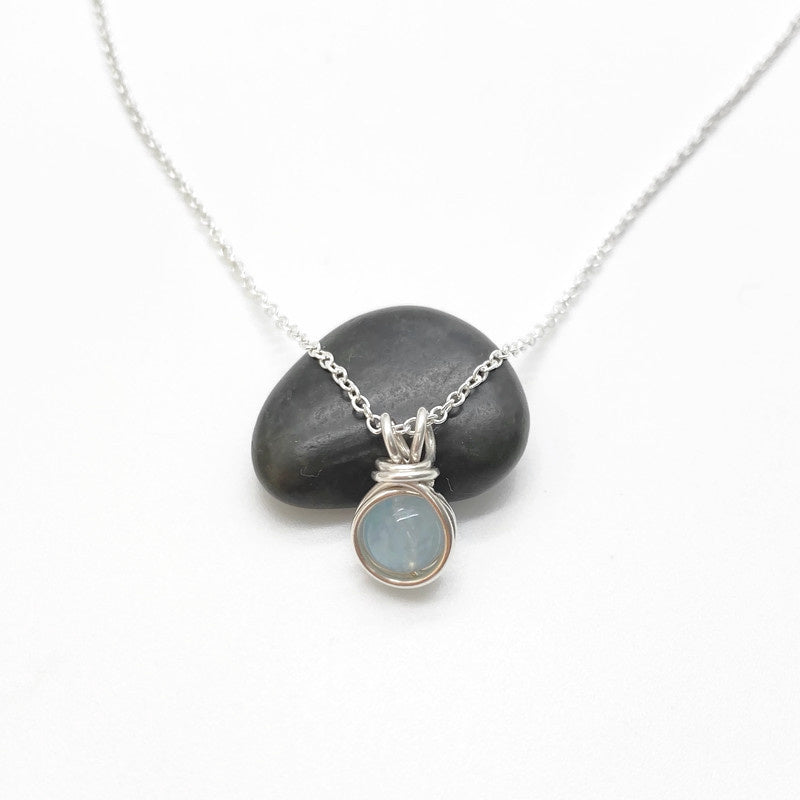 Handgemaakt zilveren collier met ronde aquamarijn hanger, gepresenteerd op een zwarte steen tegen een witte achtergrond.