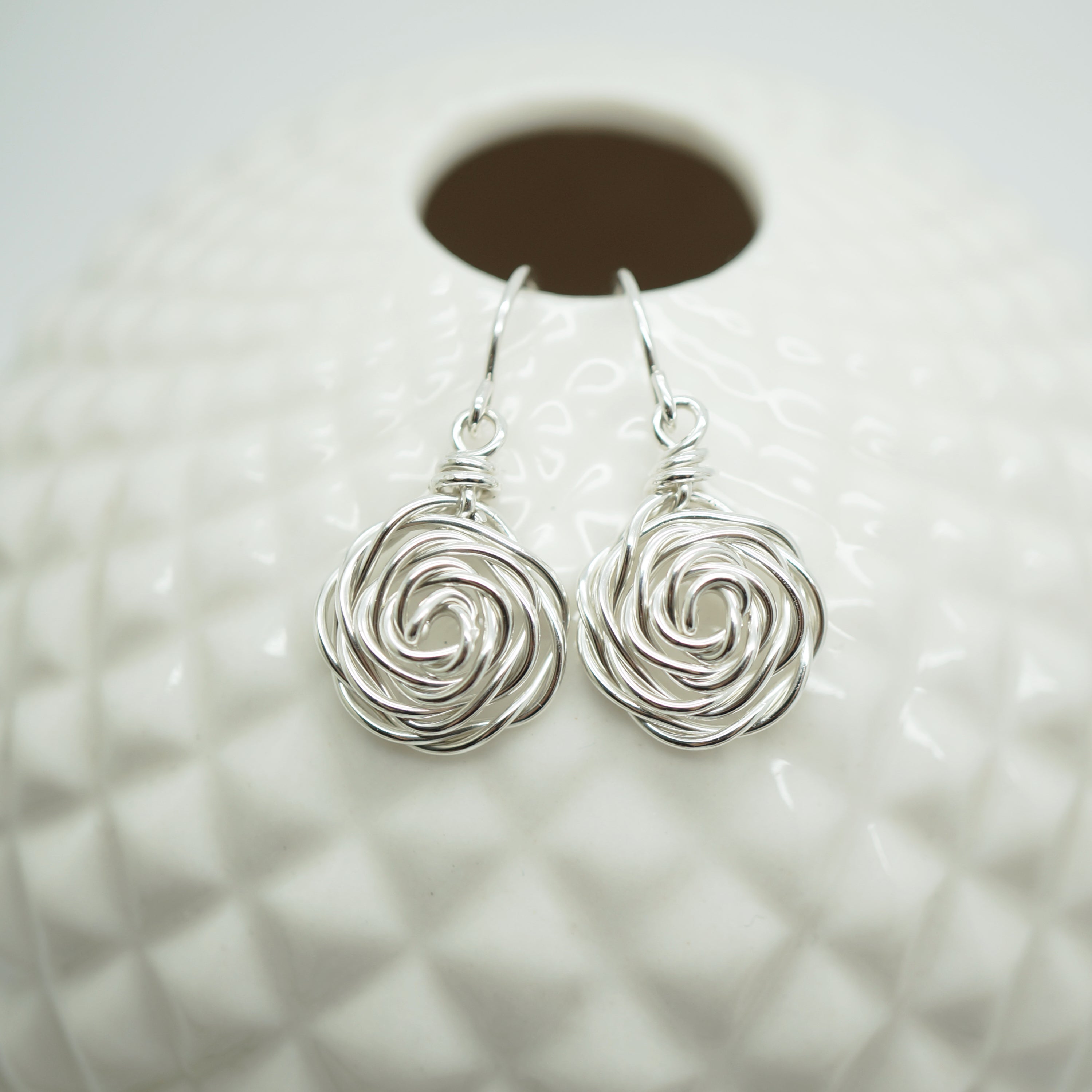 Zilveren wire wrap oorbellen in roosmotief, elegant gepresenteerd tegen een witte bolvormige achtergrond.