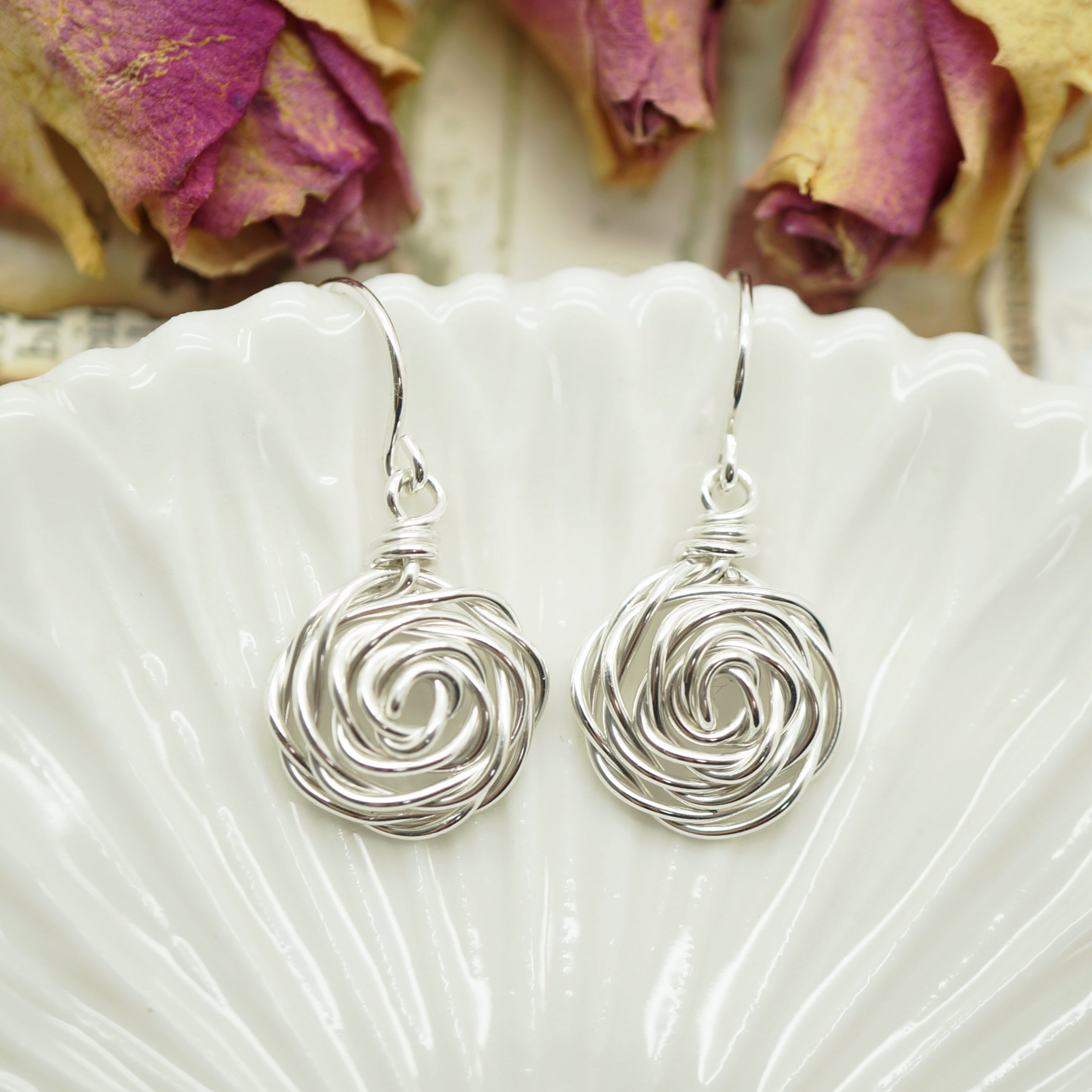 Handgemaakte roosvormige zilveren oorbellen in wire wrap stijl gepresenteerd op een wit schaaltje.
