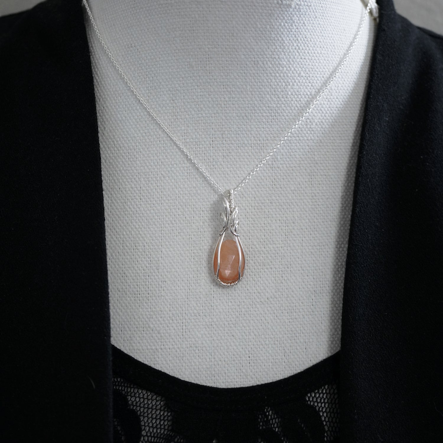 Zilveren wire wrap hanger met perzikkleurige maansteen gedragen op een buste, getoond met een zwarte blouse voor een stijlvol effect.