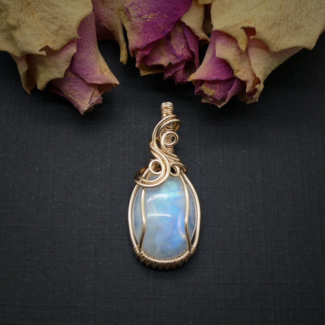 14k gold filled wire wrap hanger met regenboog maansteen, sfeervol gepresenteerd met gedroogde rozen op een donkere achtergrond