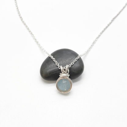 Handgemaakt zilveren collier met ronde aquamarijn hanger, gepresenteerd op een zwarte steen tegen een witte achtergrond.