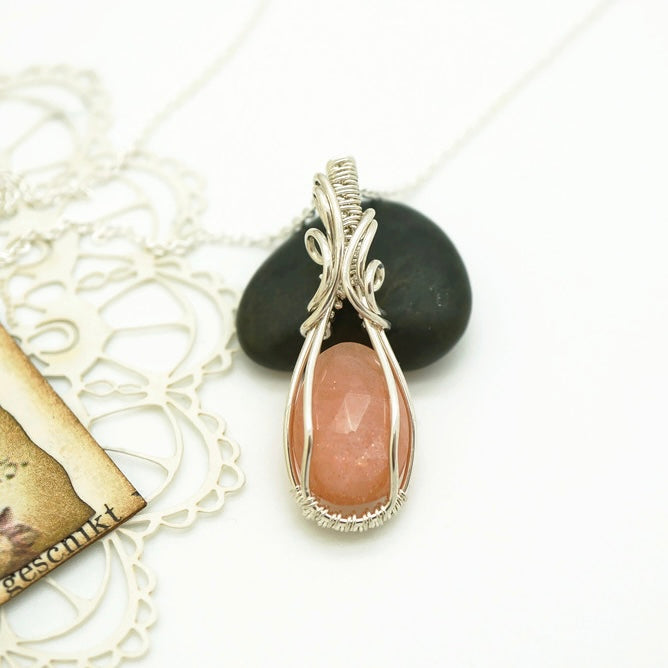 Zilveren wire wrap hanger met perzik-kleurige maansteen, handgemaakt door Atelier Blaauw, gepresenteerd op een decoratieve achtergrond.